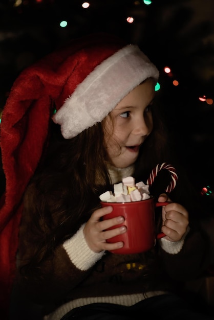 Een schattig klein meisje in een kerstmuts met een mok cacao.