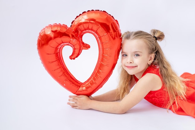 Een schattig klein meisje in een rode jurk houdt een grote ballon in de vorm van een hart op een witte geïsoleerde achtergrond het concept van Valentijnsdag een plek voor tekst