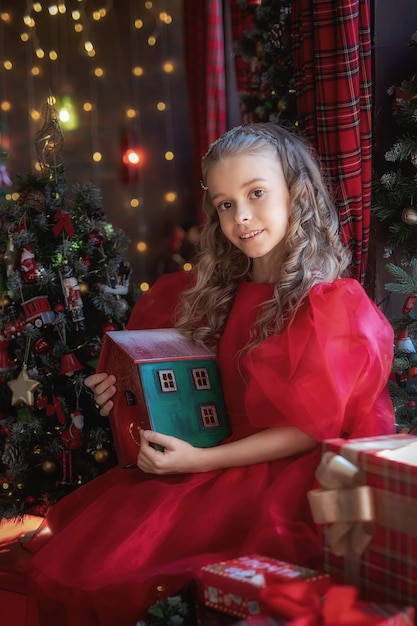 Een schattig klein meisje in een rode jurk zit bij het raam in een nieuwjaarsinterieur met een kerstboom met een houten huis in haar handen