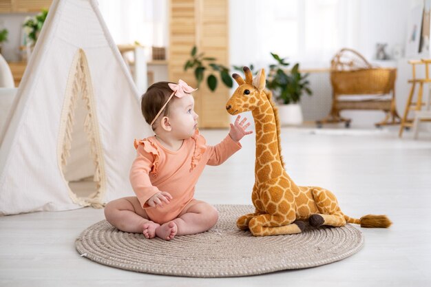 Een schattig klein meisje in een roze bodysuit zit op een kleed in de woonkamer van het huis tegen de achtergrond van een wigwam die speelt met een opgezette giraffe