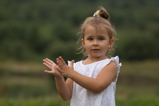 Een schattig klein meisje in een witte jurk staat op een zomeravond aan de oever van een meer en klapt in haar handen Zonsondergang op het meer