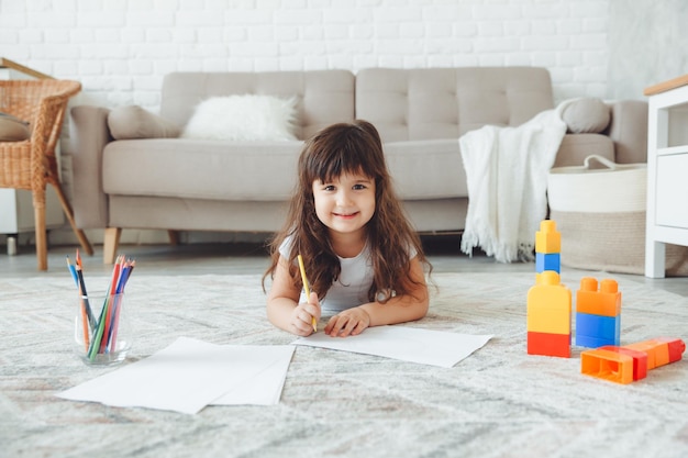 Een schattig klein meisje ligt op de vloer van het huis en tekent met potloden voor de vrije tijd van kinderen