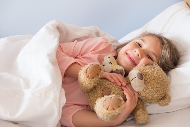 Een schattig klein meisje met blond haar in een roze t-shirt ligt in bed glimlachend en knuffelend een teddybeer