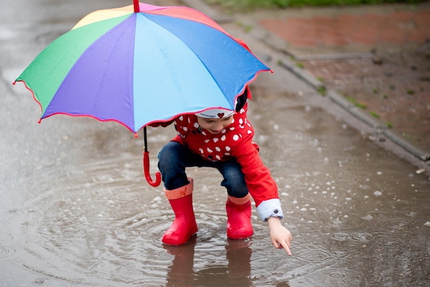 Een schattig klein meisje springt in plassen en heeft plezier Het meisje heeft een regenboogparaplu in haar handen