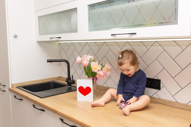 Een schattig klein meisje zit in de keuken met een boeket roze tulpen mooie cadeau ansichtkaart