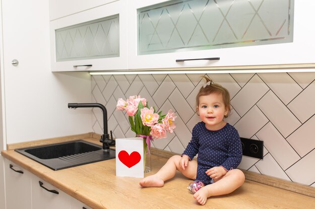 Een schattig klein meisje zit in de keuken met een boeket roze tulpen mooie cadeau ansichtkaart