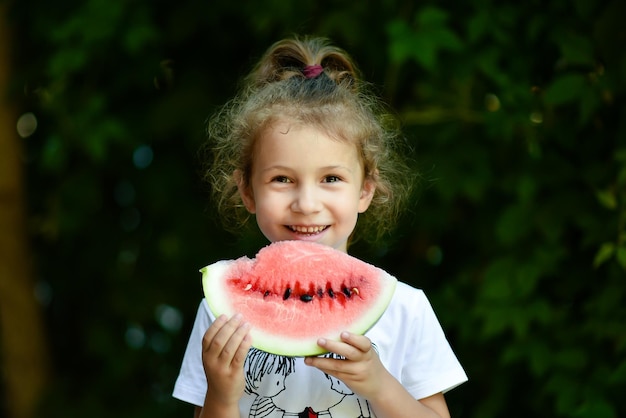 Een schattig lachend meisje eet een rode watermeloen Achtergrond van groene bladeren Nature Food Summer