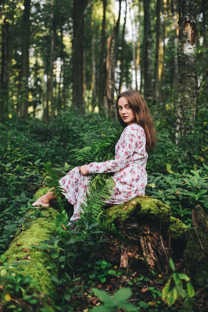 Een schattig meisje in een bloemenjurk zit met een varenboeket in het bos.