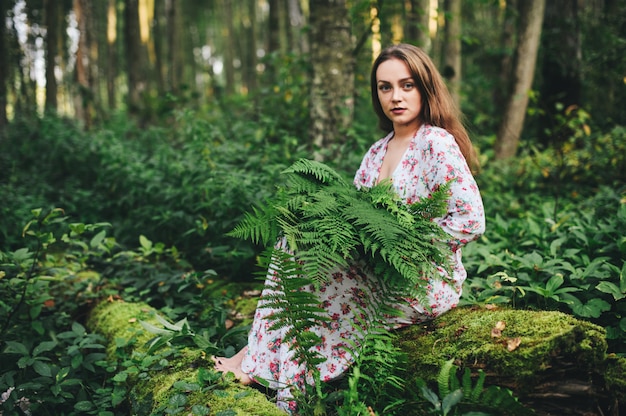 Een schattig meisje in een bloemenjurk zit met een varenboeket in het bos.