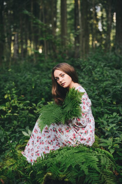 Een schattig meisje in een bloemenjurk zit met een varenboeket in het bos.