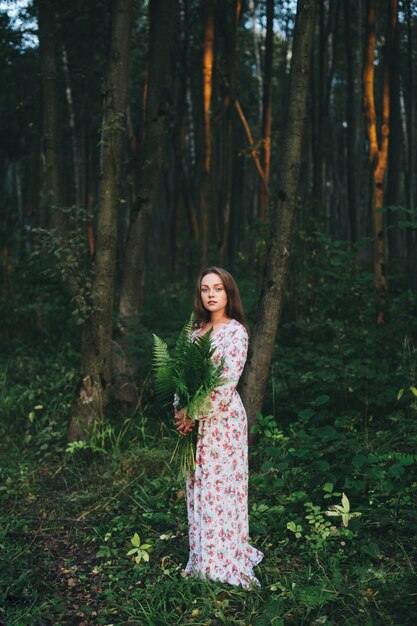 Een schattig meisje in een bloemenjurk zit met een varenboeket in het bos.