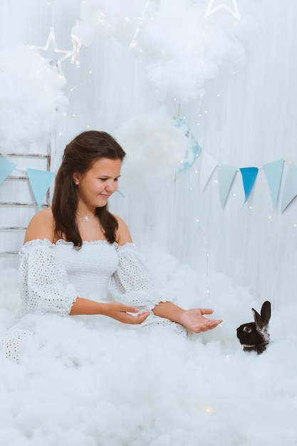 Een schattig meisje in een witte outfit zit in de wolken en strekt haar handen uit naar een zwart konijn