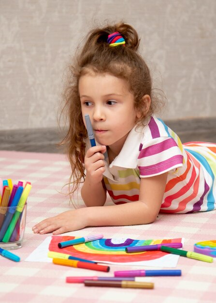 Een schattig meisje in regenboogkleren tekent een regenboog met veelkleurige stiften House Childhood