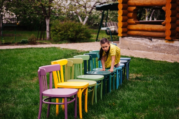 Een schattig meisje kruipt op haar knieën op de stoelen van kleurrijke Weense houten stoelen op een groen gazon in de buurt van...