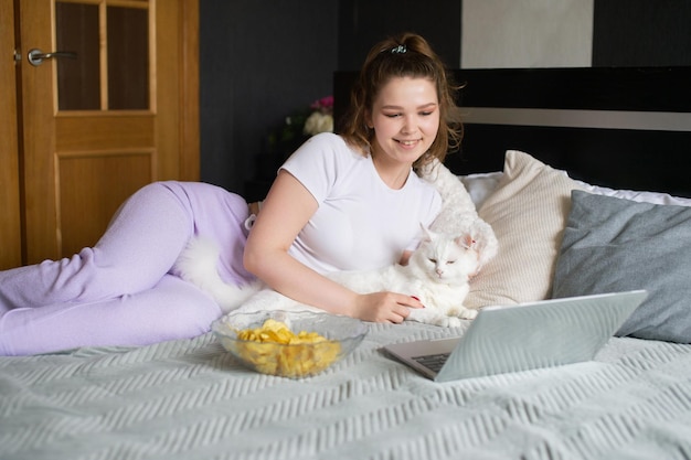 Een schattig meisje ligt op het bed en aait de kat en kijkt naar haar laptopchips in een bord