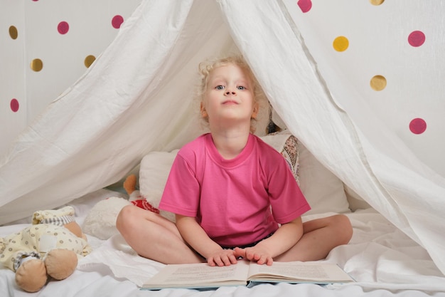 Een schattig meisje met krullend haar leest een boek in een hut van een laken op het bed, een klein meisje dat plezier heeft en speelt in haar tent, een hut in de kinderkamer