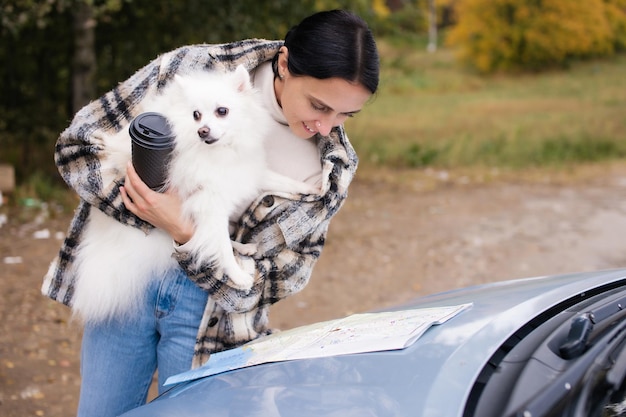 Een schattig meisje staat bij de auto met een witte Pommerse Pommeren en een koffiekopje in haar handen