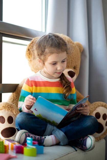 Een schattig meisje van 6 jaar zit op de bank bij het raam met een grote speelgoedbeer en leest een boek