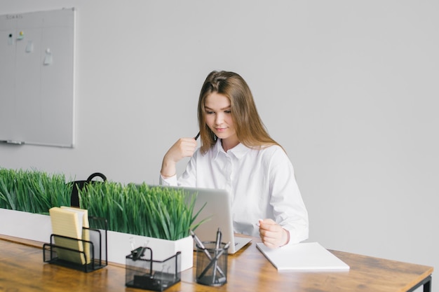 Een schattig meisje zit aan een houten bureau in het kantoor en werkt met een laptop en notepad