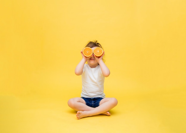 Een schattig meisje zit en houdt de helften sinaasappelen voor haar ogen. een klein meisje zit in kleermakerszit. een klein meisje in een wit t-shirt en denim shorts op een gele ruimte.