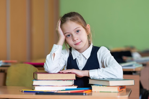 Een schattig schoolmeisje zit aan een bureau omringd door boeken Kijkt in de camera Schoolleven