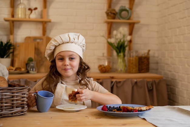 een schattig zesjarig meisje met een koksmuts en schort zit aan een houten tafel pannenkoeken te eten