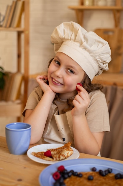 een schattig zesjarig meisje met een koksmuts en schort zit aan een houten tafel pannenkoeken te eten