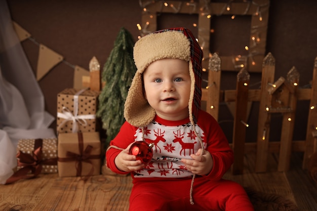 een schattige baby in oudejaarspyjama en een muts met oorkleppen houdt een rode nieuwjaarsbal vast