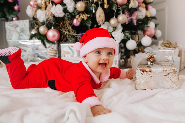 Een schattige baby van 6 maanden oud in een kerstmankostuum ligt op een deken bij de kerstboom. kerst 2022