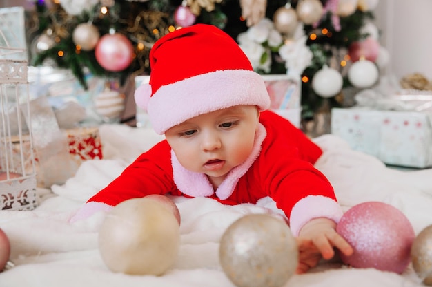 Een schattige baby van 6 maanden oud in een kerstmankostuum ligt op een deken bij de kerstboom. kerst 2022