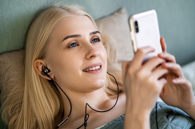 Een schattige blondine met blauwe ogen ligt op het bed. In haar hand heeft ze een telefoon waarop een zwarte koptelefoon is aangesloten.