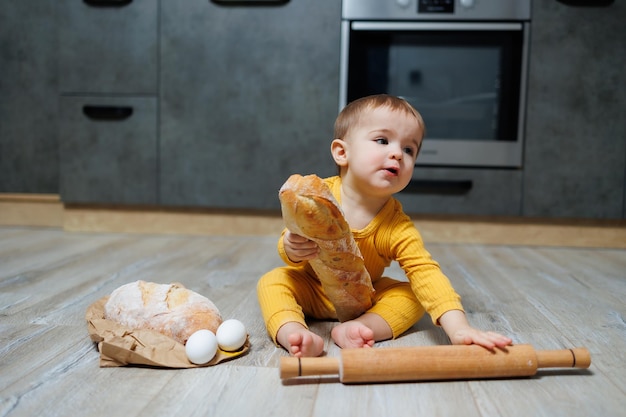 Een schattige eenjarige jongen zit in de keuken en eet een lang brood of stokbrood in de keuken Het eerste brood eten door een kind Brood is goed voor kinderen