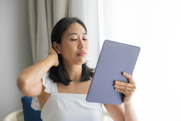 Een schattige, gelukkige aziatische vrouw die een witte jurk draagt, speelt met zijn tablet op een crèmekleurige bank en kijkt naar het mobiele scherm