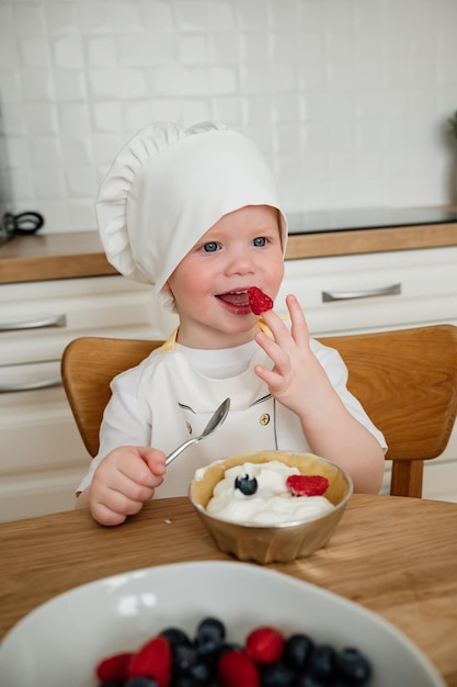Foto een schattige gelukkige jongen met een chefhoed en schort die frambozen van zijn vingers eet.