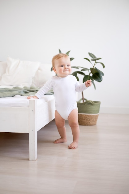 Een schattige gezonde babyjongen in een ruime lichte slaapkamer staat naast een bed met wit linnengoed het kind staat met steun het kind leert lopen