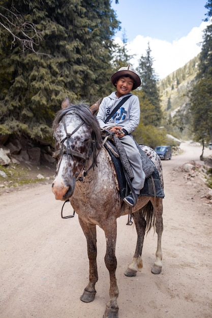 Een schattige, glimlachende aziatische jongen met een nationale hoed die op een paard rijdt, rijdt in de bergen en ontmoet toeristen