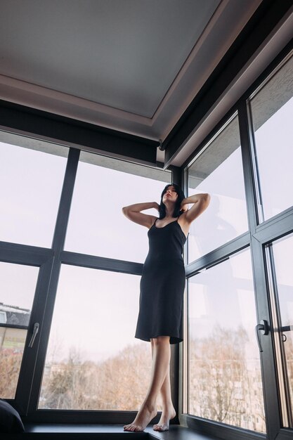 Een schattige gracieuze brunette in een lichtzwarte jurk op de achtergrond van een panoramisch raam 4559