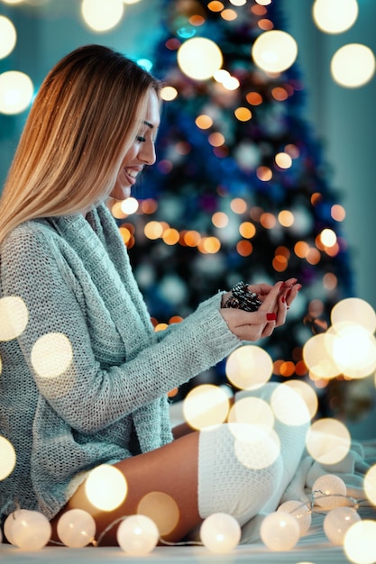 Een schattige jonge lachende vrouw houdt dennenappels in haar handen omringd met kerstverlichting.