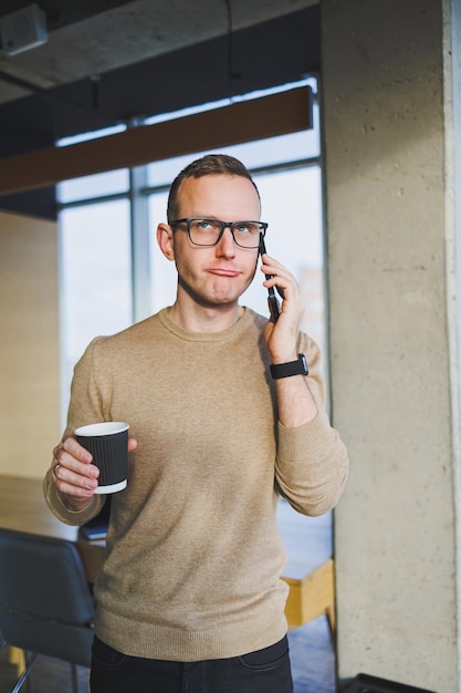 Een schattige jonge man in een bruine trui en bril praat emotioneel op een mobiele telefoon en drinkt koffie terwijl hij ontspant op kantoor Een jonge freelancer werkt op afstand