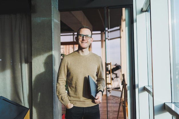 Een schattige jonge man in een bruine trui en bril staat bij een groot raam op kantoor met een laptop in zijn handen terwijl hij op kantoor werkt Een jonge freelancer werkt op afstand