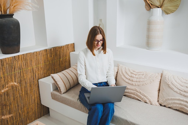 Een schattige jonge vrouw in casual kleding zit thuis op de bank en werkt aan een laptop. Werk online op afstand.