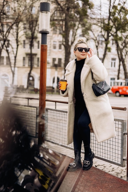 Een schattige jonge vrouw in een witte bontjas en een kopje koffie loopt door de stad Stijlvol jong zwanger meisje Casual mode voor dames