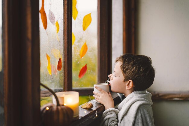 Een schattige jongen gewikkeld in een deken drinkt hete thee en kijkt uit het open raam naar de prachtige herfstnatuur Herfst interieur Gezellige herfststemming Thanksgiving Halloween