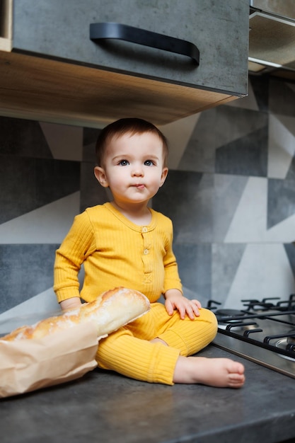 Een schattige jongen van 1 jaar oud in gele kleren zit in de keuken op tafel met een stokbrood van verse tarwe Portret van een schattige jongen van een jaar die heerlijk brood eet