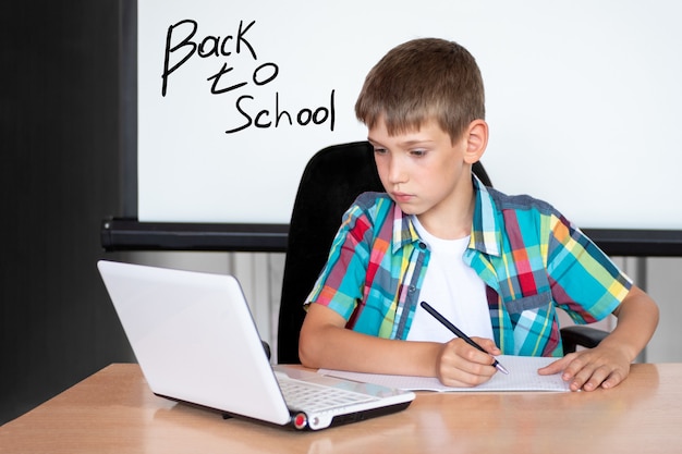 Een schattige jongen zit aan een tafel, kijkt naar een laptop, schrijft huiswerk of bereidt zich voor op een examen. Concept Terug naar school. Ruimte kopiëren