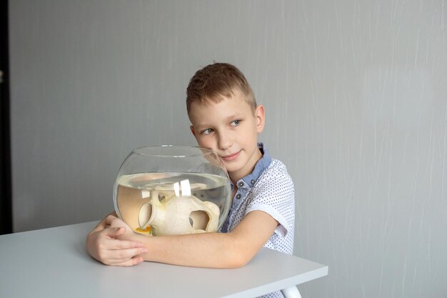 Een schattige jongen zit bij een transparant aquarium in de kamer en knuffelt een aquarium met vissen