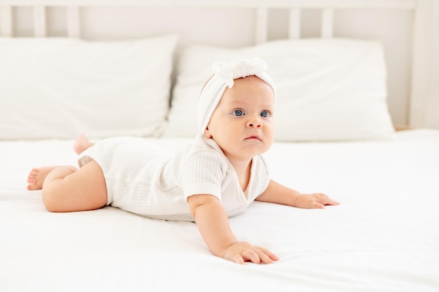 Een schattige kleine baby in een witte bodysuit ligt op zijn buik op een bed in een lichte slaapkamer op een katoenen bed een pasgeboren babymeisje met blauwe ogen, gezonde en gelukkige glimlach