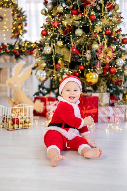 Een schattige kleine babyjongen in een kerstmankostuum zit met kerstboomspeelgoed onder een feestelijke kerstboom van het huis een gelukkig kind viert thuis Kerstmis en Nieuwjaar