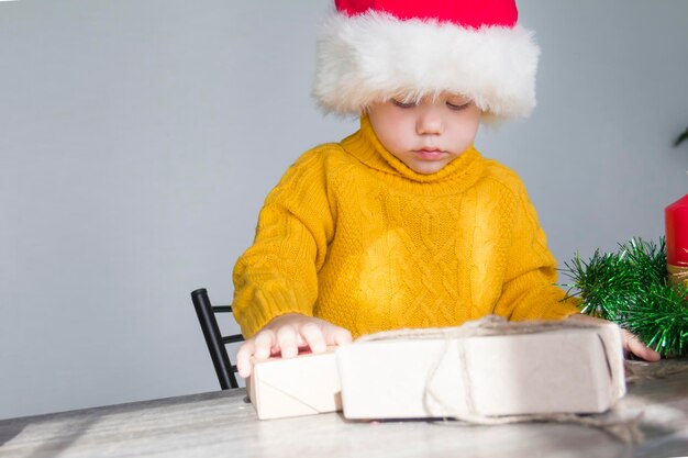 Een schattige kleine jongen in een gele trui en een rode kerstmuts aan een houten tafel met geschenken die er zijn