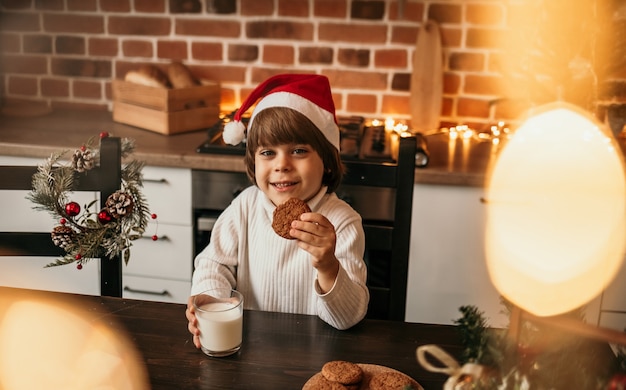 Een schattige kleine jongen in een witte gebreide trui en een rode kerstmuts zit aan de keukentafel en eet havermoutkoekjes en melk